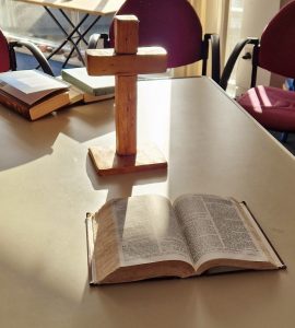 A bible resting open in front of a small 1 foot cross on top of a pale coloured table.
